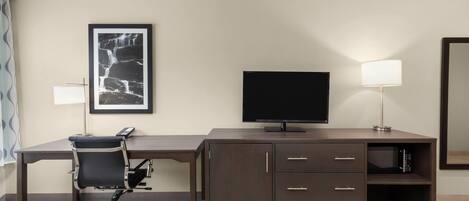 A hotel room with a dark wooden desk, a black office chair, a flat-screen TV, a lamp, a framed picture on the wall, and a mirror.