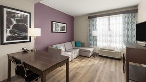 Une chambre d’hôtel moderne avec un bureau en bois, un canapé, une télévision et une grande fenêtre agrémentée de rideaux à motifs.