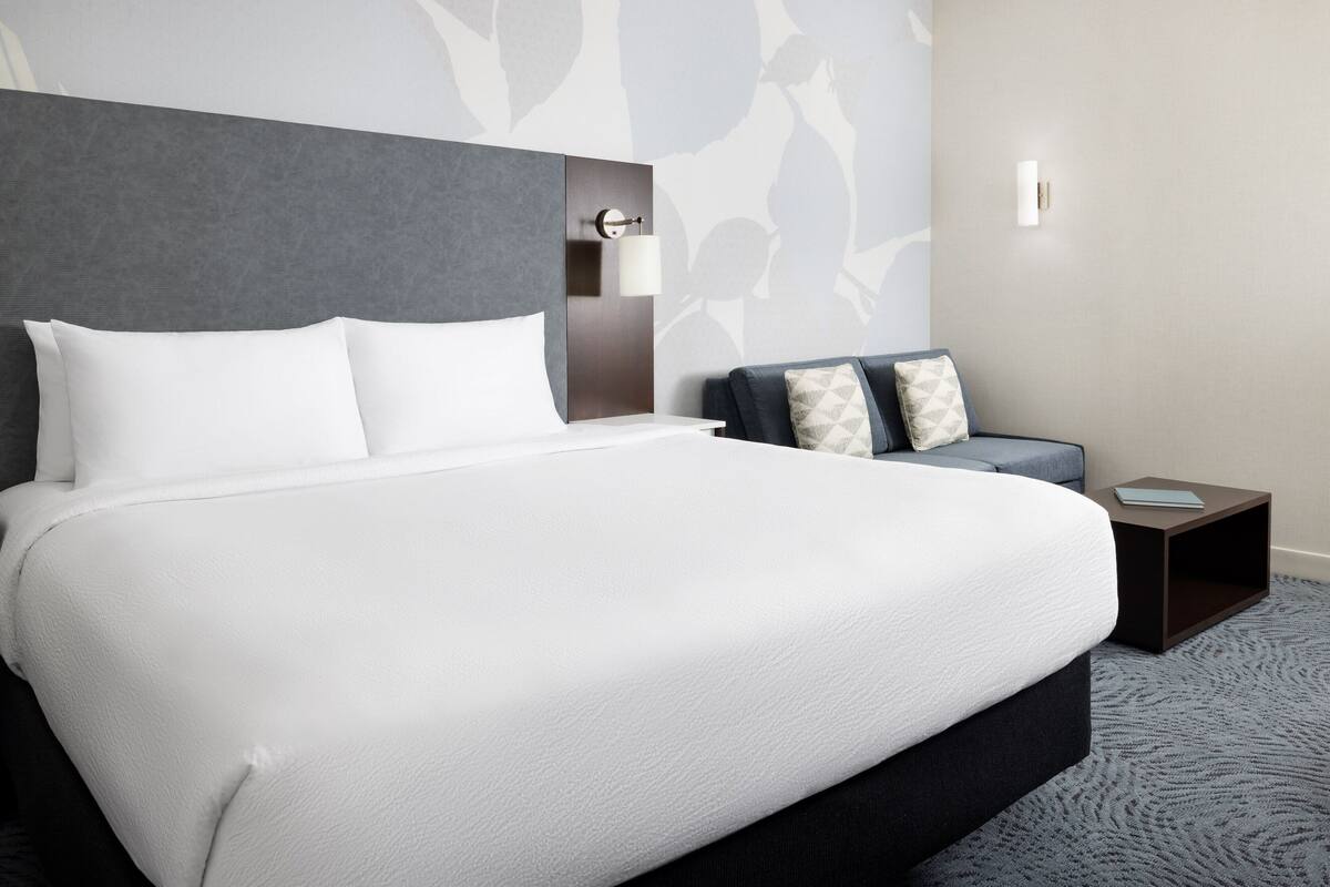 Una habitación de hotel moderna con una cama grande, un cabecero gris y una mesita de noche pequeña con una lámpara.