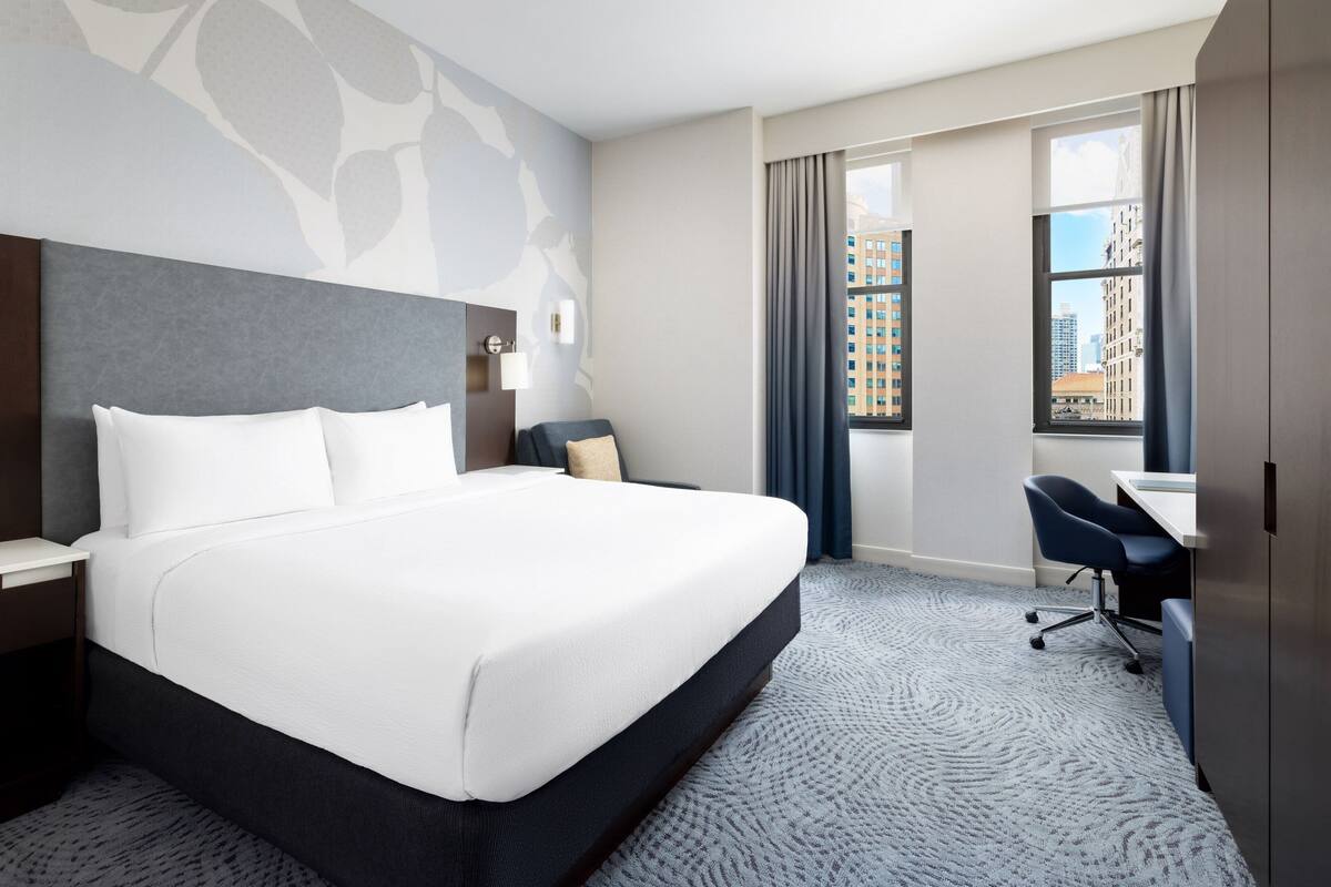 Habitación de hotel con una cama grande, un escritorio, una silla y vista a la ciudad desde la ventana.