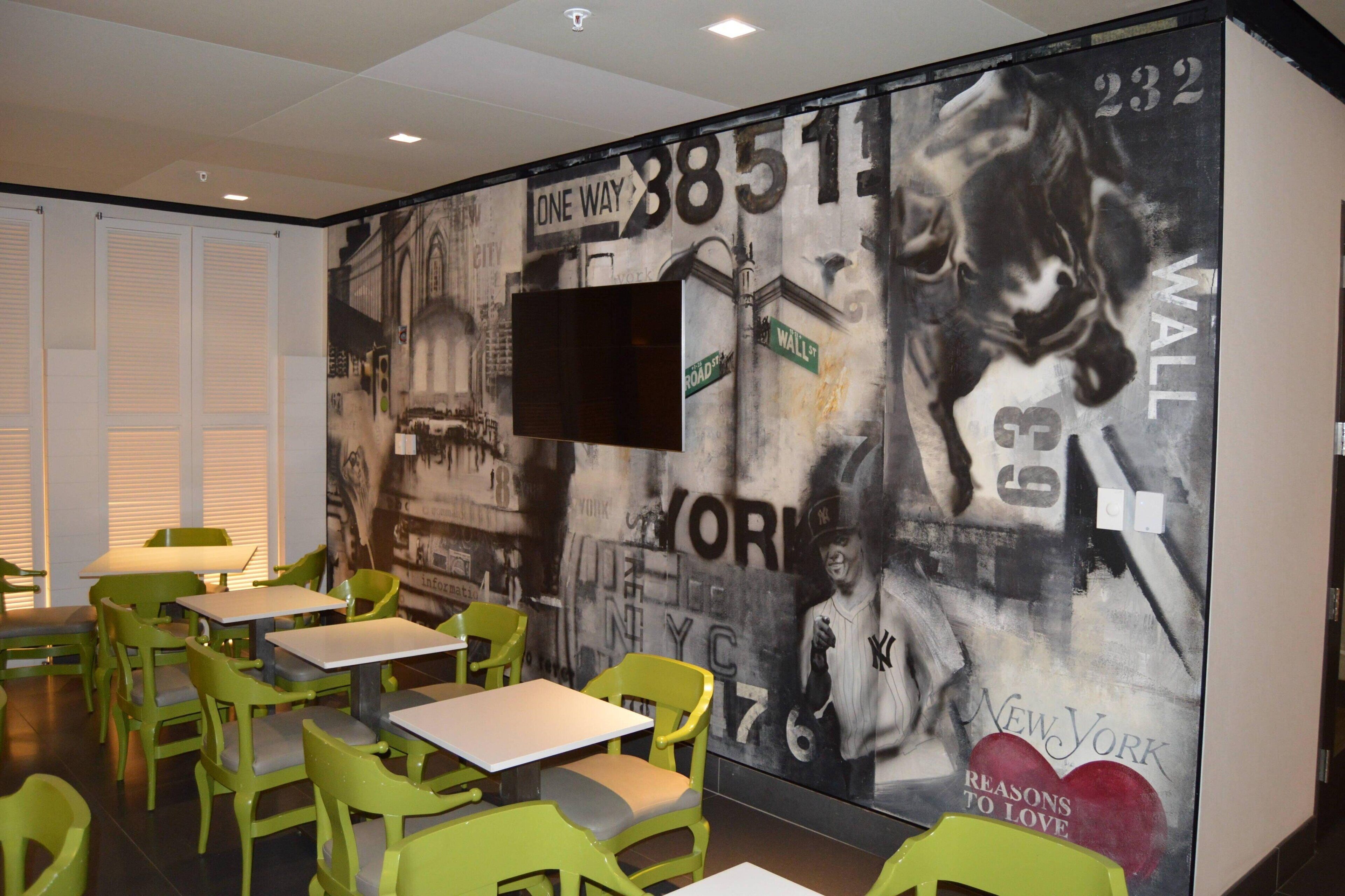 A restaurant with green chairs and tables, a mural with cityscape and sports imagery, and a mounted TV.