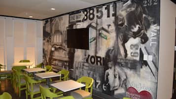 A restaurant with green chairs and tables, a mural with cityscape and sports imagery, and a mounted TV.