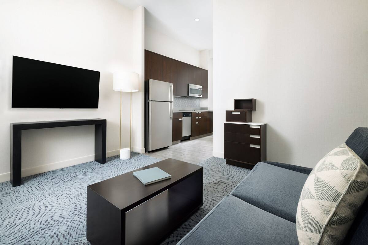 Una habitación de hotel moderna con una televisión de pantalla plana, un sofá, una mesita de centro, una zona de cocina con refrigerador y microondas, y una lámpara de noche.