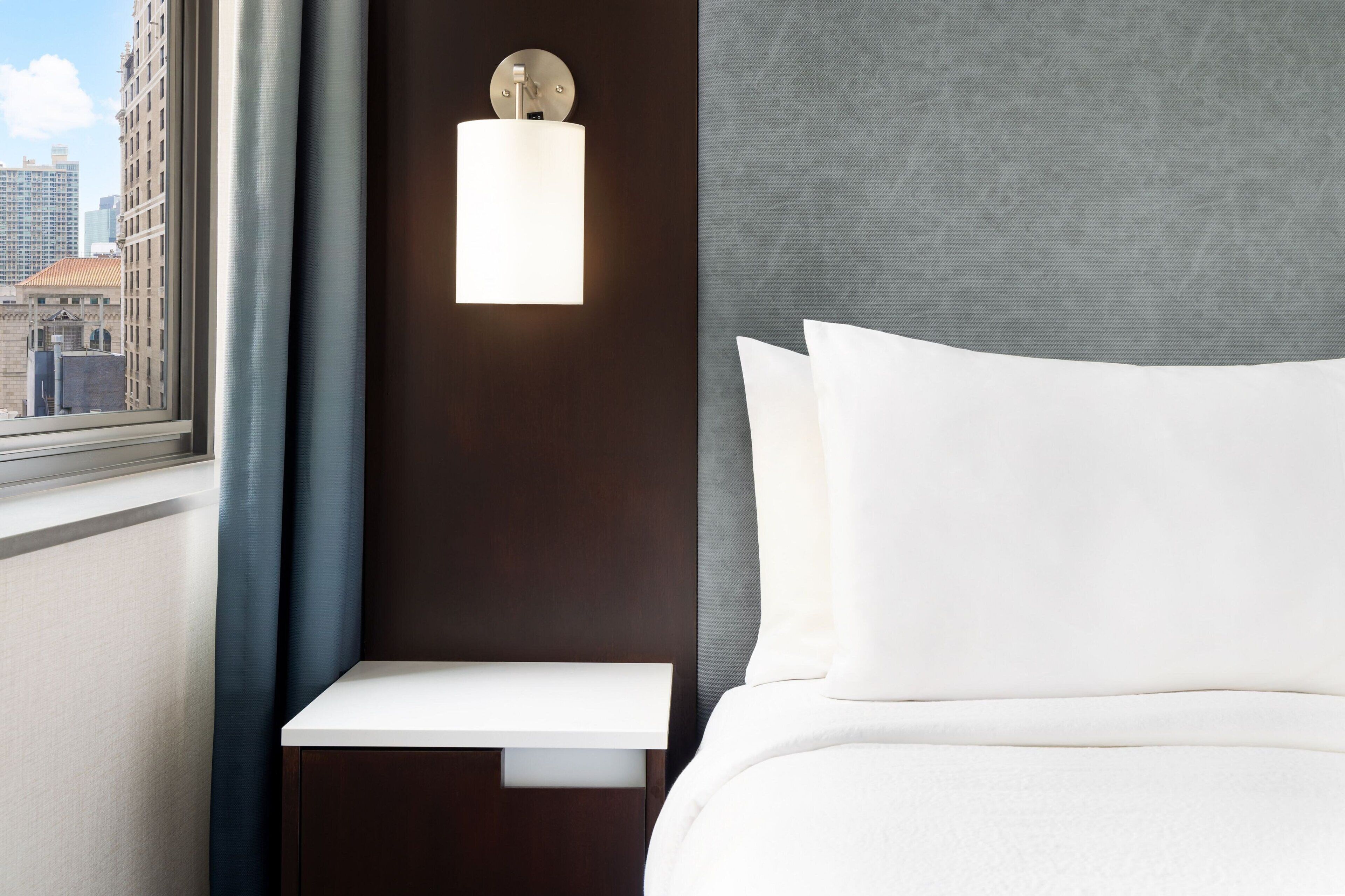 A hotel room with a large window, a bedside table, a wall-mounted light fixture, and a bed with white bedding.