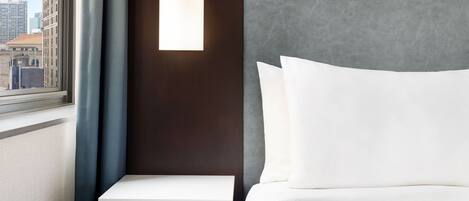 Habitación de hotel con un ventanal grande, una mesita de noche, una lámpara de pared y una cama con ropa blanca.