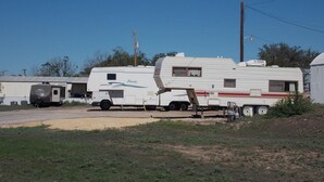 A white recreational vehicle with a sliding door and a small attached structure, parked in a rural area with a dirt ground and sparse vegetation. - Holiday Host Motel (Sonora)