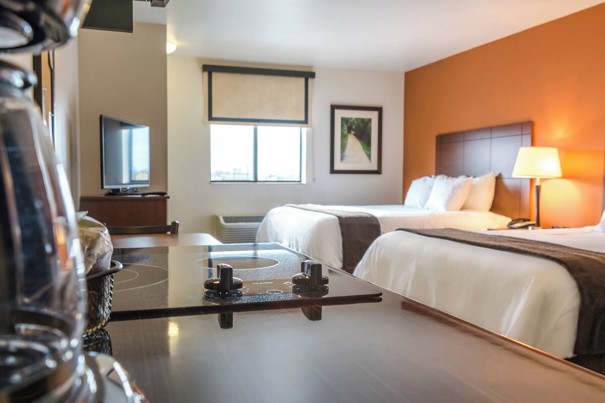 A hotel room with two beds, a TV, a window with blinds, a framed picture on the wall, and a bedside lamp.