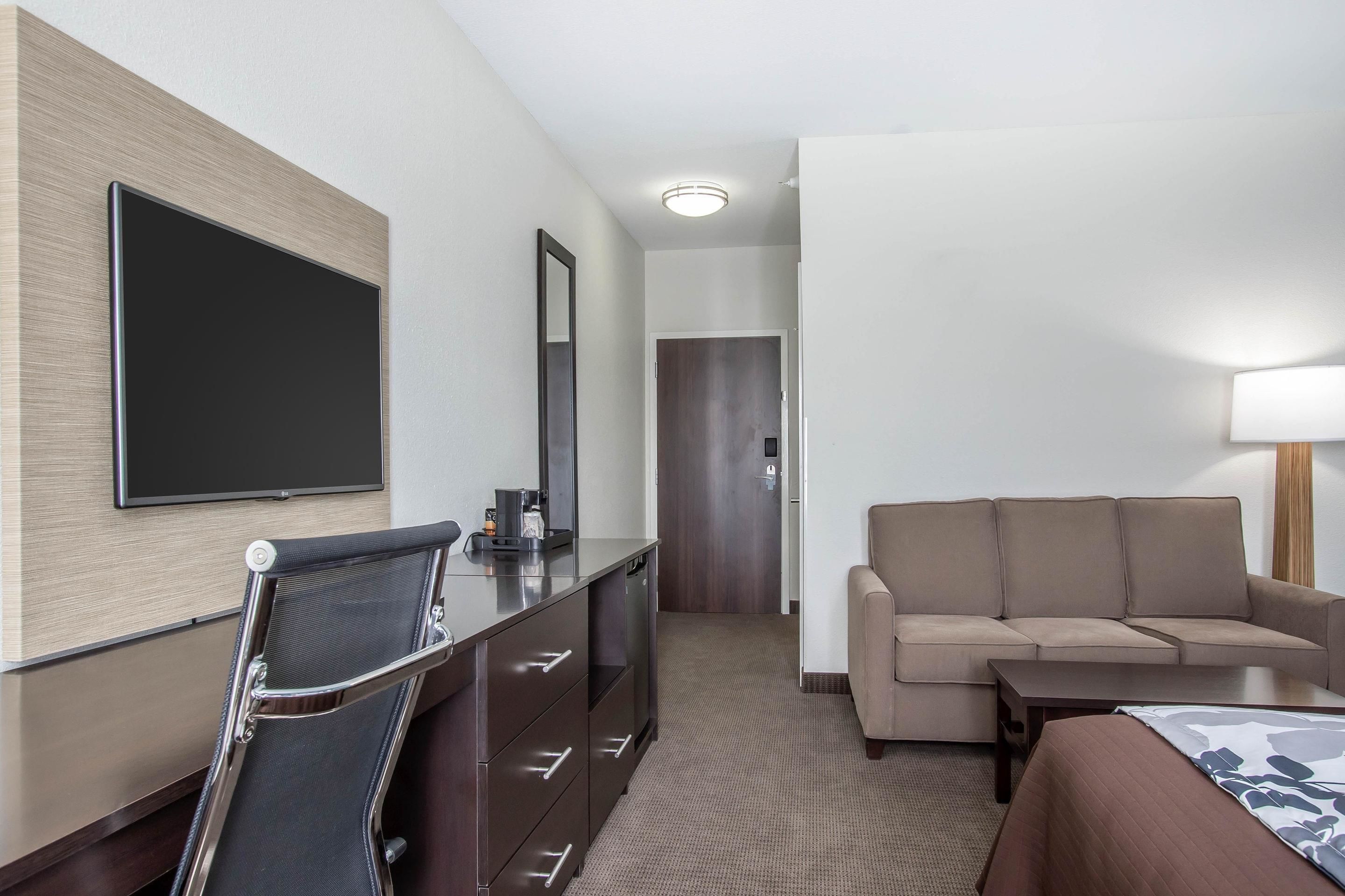 A hotel room with a bed, a sofa, a desk with a chair, a flat-screen TV, and a mirror.
