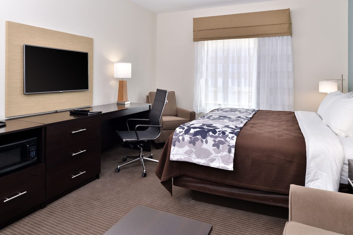A hotel room with a bed, a desk with a chair, a flat-screen TV mounted on the wall, a microwave, and a window with curtains.