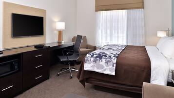 A hotel room with a bed, a desk with a chair, a flat-screen TV mounted on the wall, a microwave, and a window with curtains.