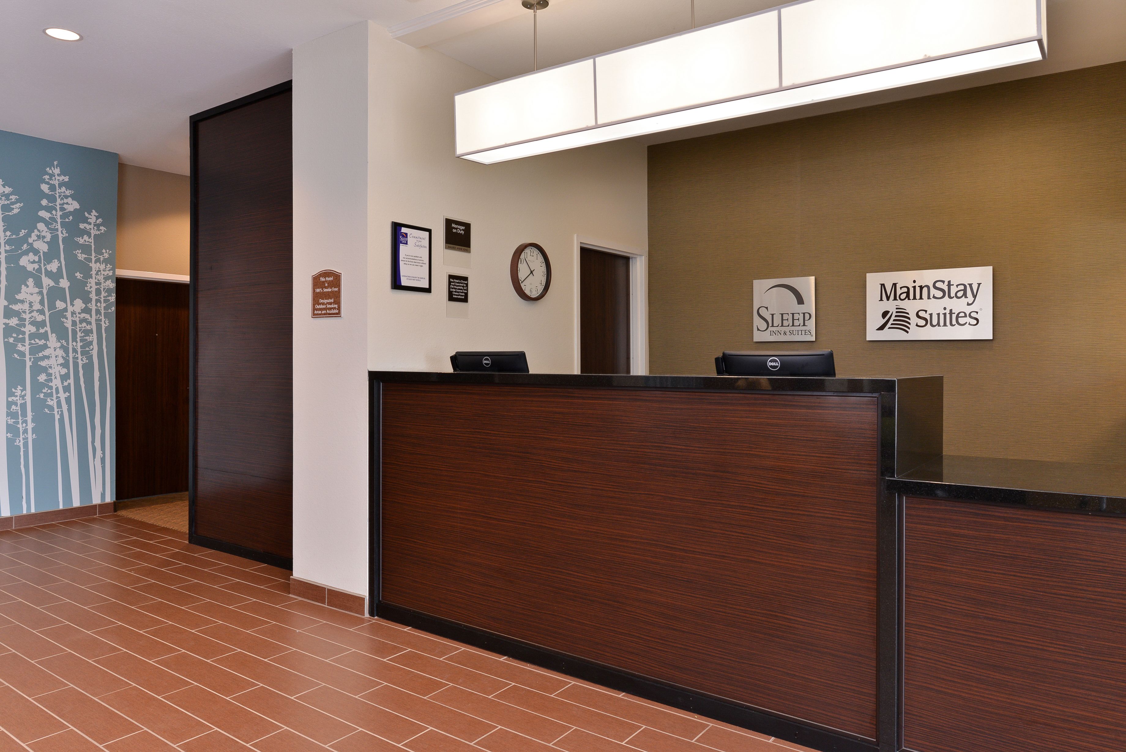 A hotel reception area with a wooden reception desk, a clock, and signs for 'Sleep Inn & Suites' and 'MainStay Suites'.