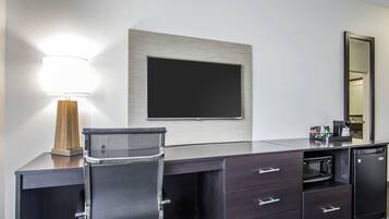 Habitación de hotel con escritorio, una silla, televisor de pantalla plana, espejo, lámpara, microondas y refrigerador.