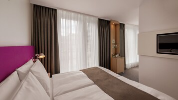 Una habitación de hotel con cama, escritorio, televisión y ventana con cortinas.