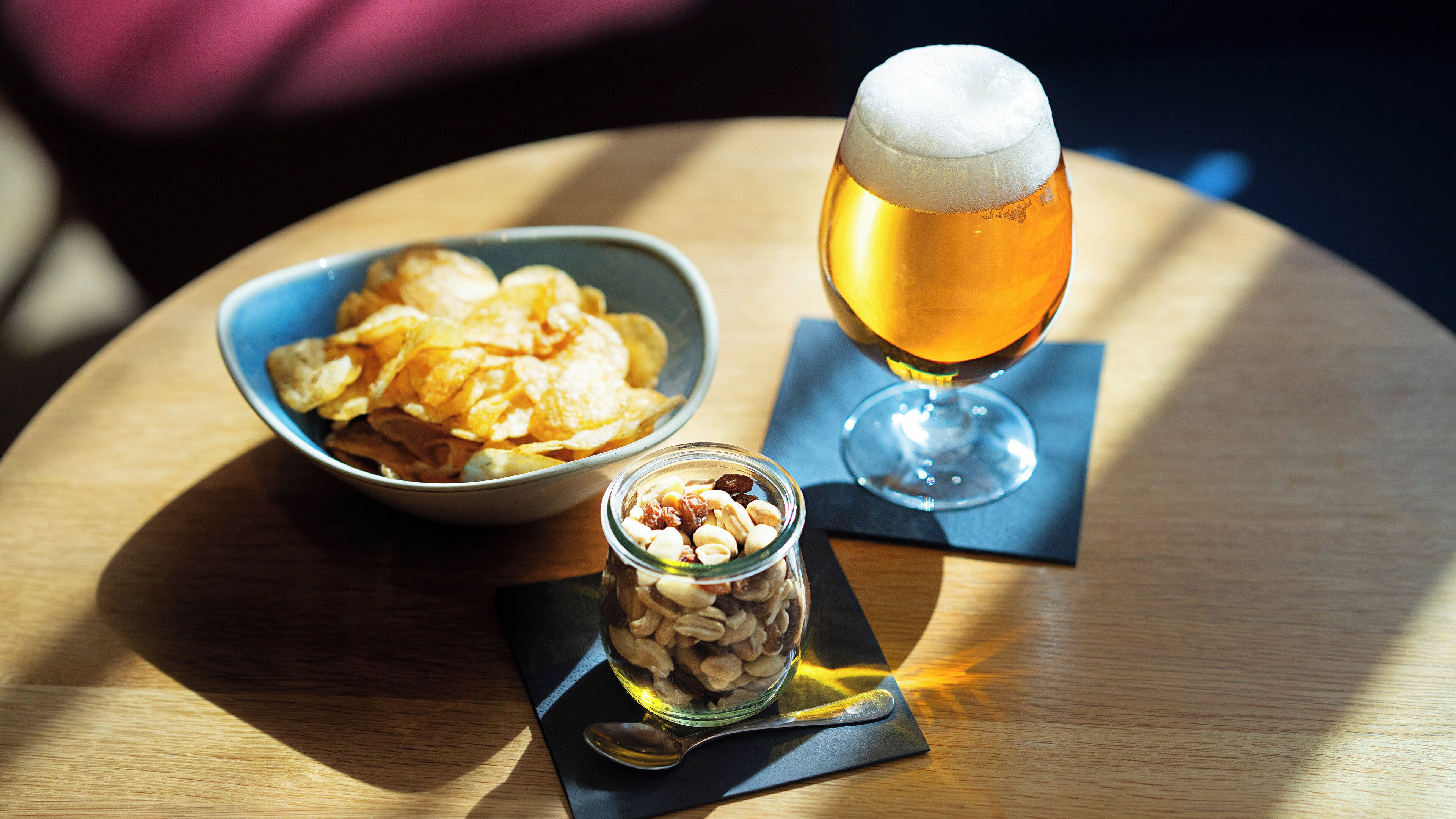 A bowl of chips, a glass of beer, and a jar of nuts on a wooden table.