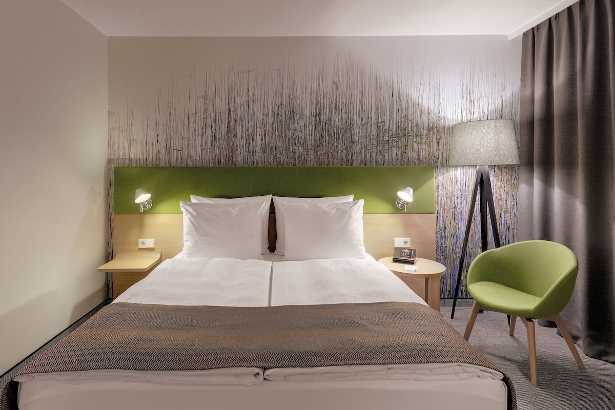 A hotel room with a large bed, two bedside tables, a green chair, and a decorative wall panel.