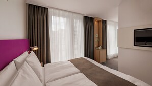 Ein Hotelzimmer mit einem Bett, einem Schreibtisch, einem Fernseher und großen Fenstern mit Vorhängen.