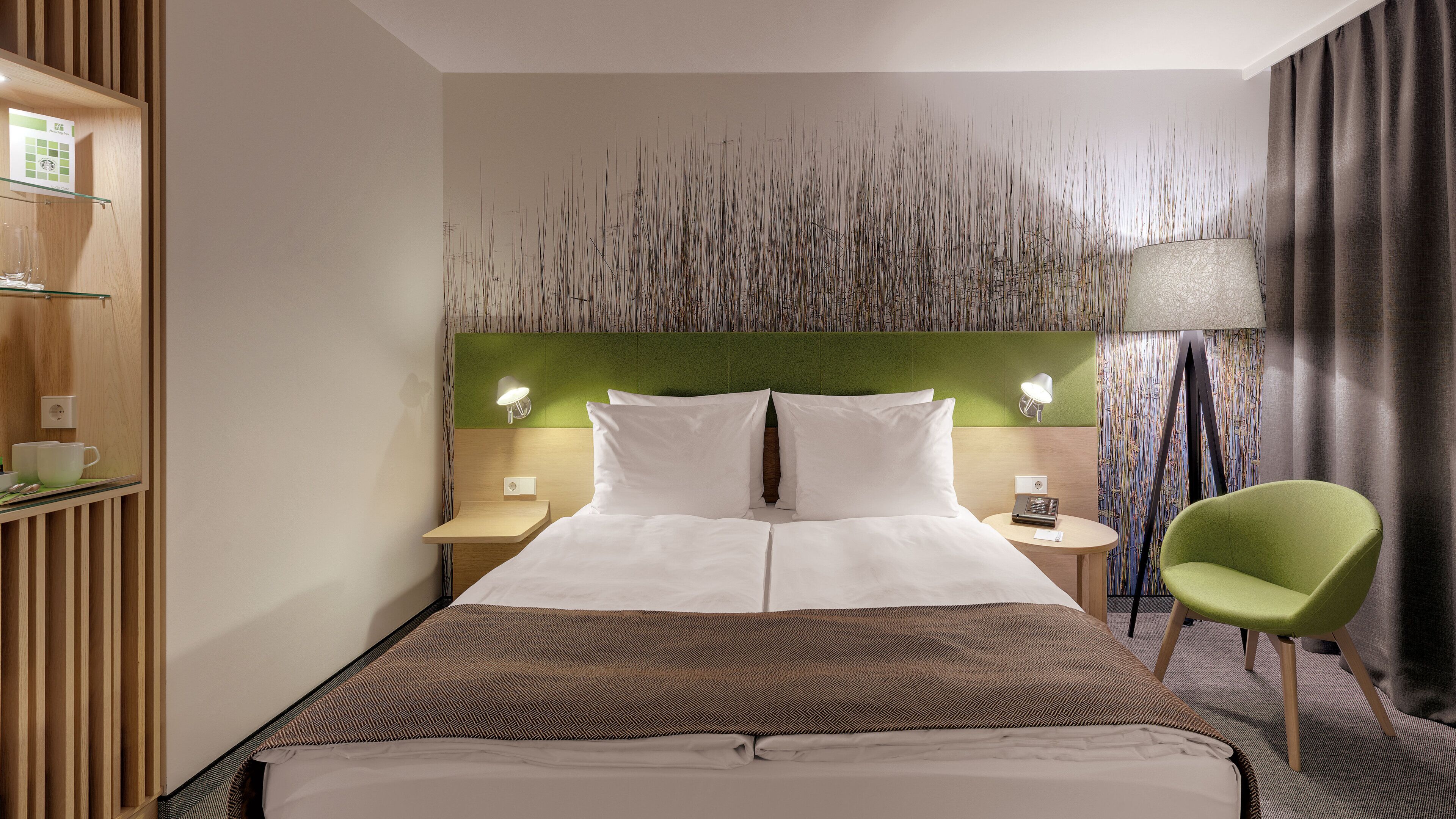 A hotel room with a bed, two bedside tables, a green chair, and a decorative wall panel.