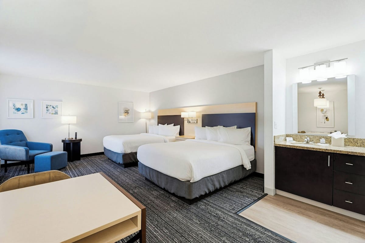 A hotel room with two beds, a desk, a chair, a bathroom with a mirror and sink, and artwork on the walls.