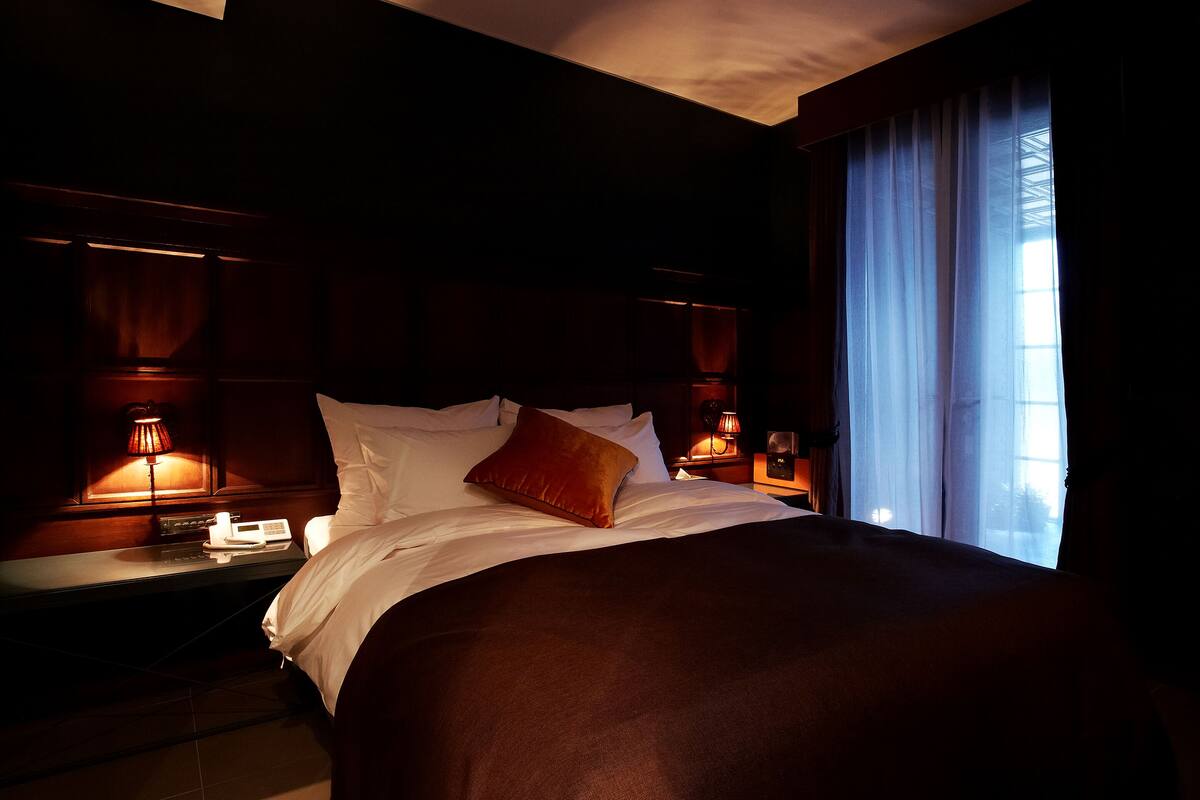 Una habitación de hotel con una cama grande, dos mesitas de noche con lámparas y una ventana con cortinas.