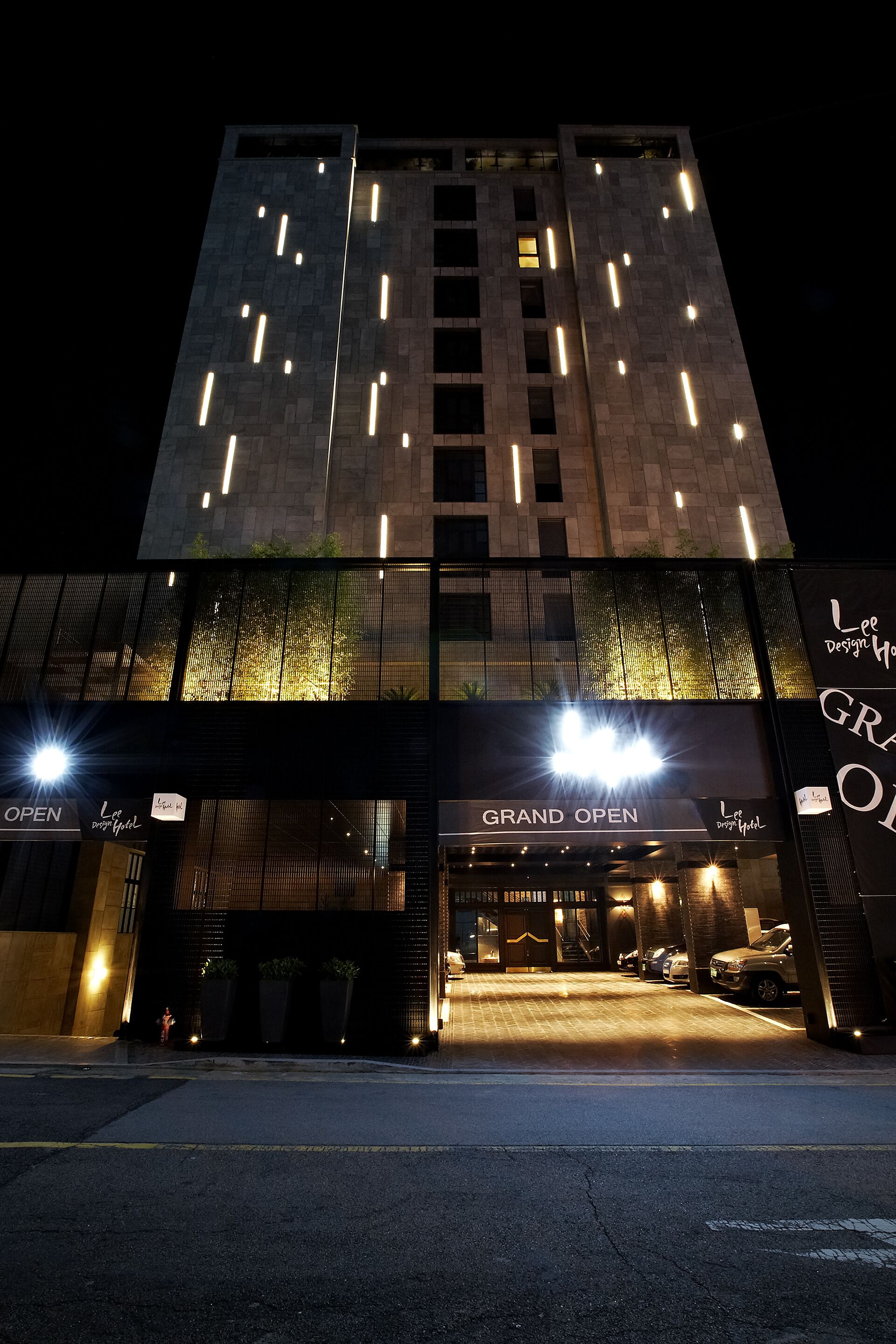 A modern building with a 'Grand Open' sign, illuminated entrance, and parked cars.