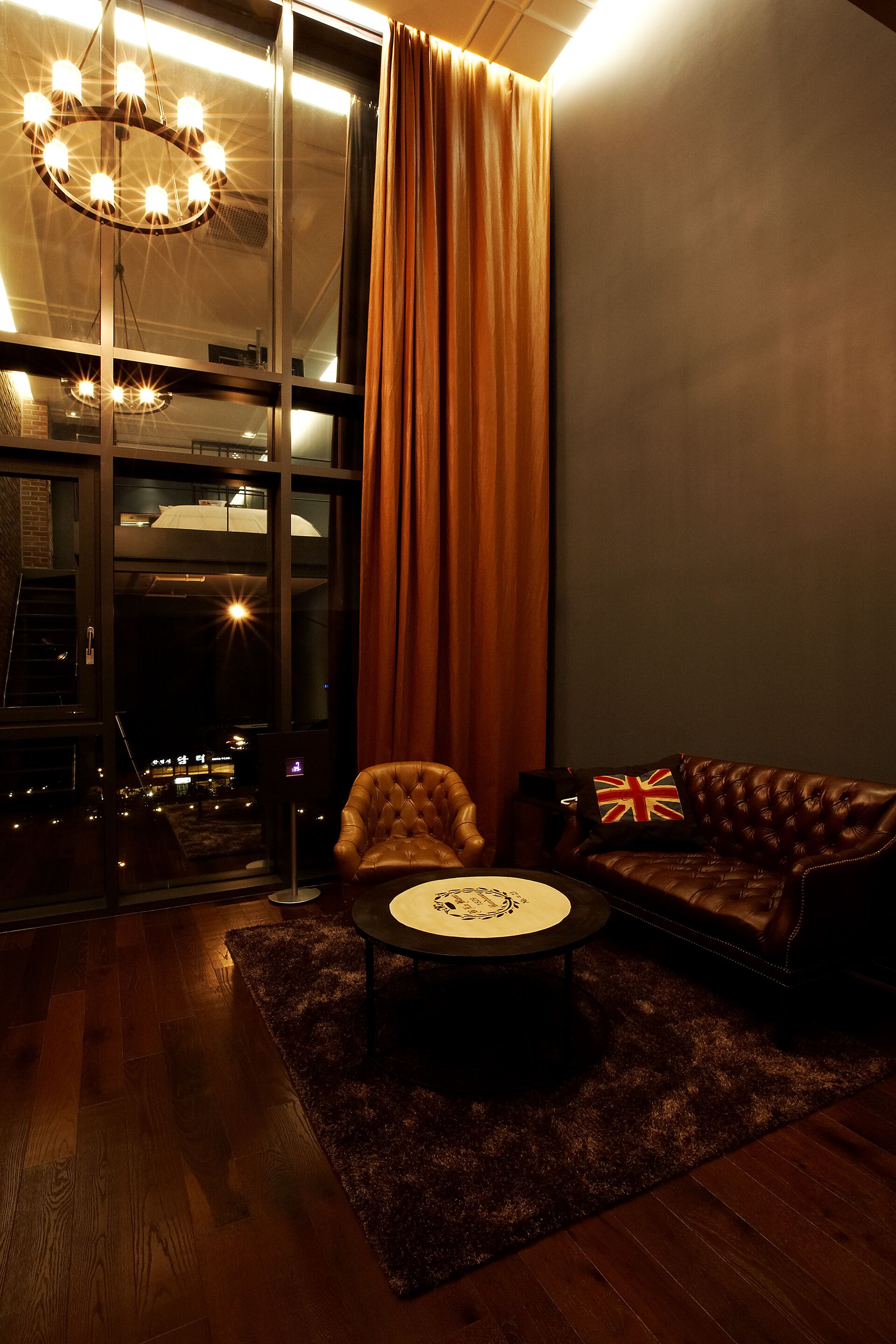 A cozy living room with a leather sofa, a tufted armchair, and a round coffee table. A large window with curtains allows natural light and views of the city.