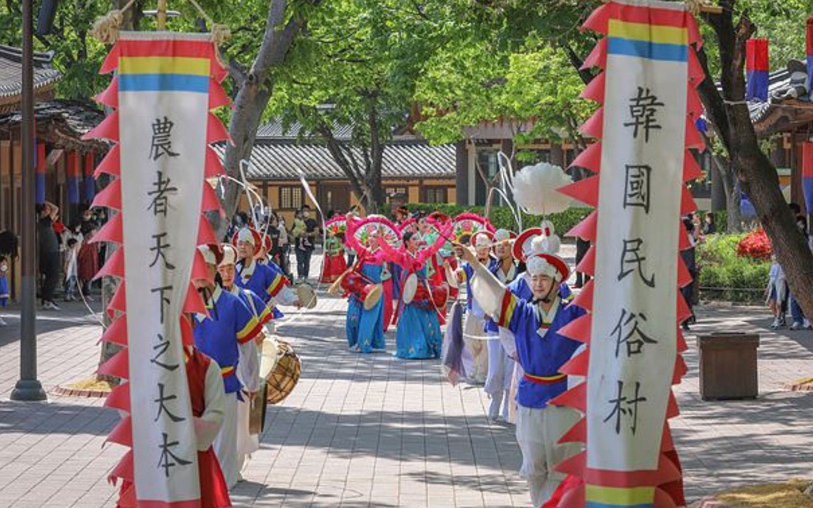A traditional parade with performers in colorful costumes and fans, walking down a paved street with banners.
