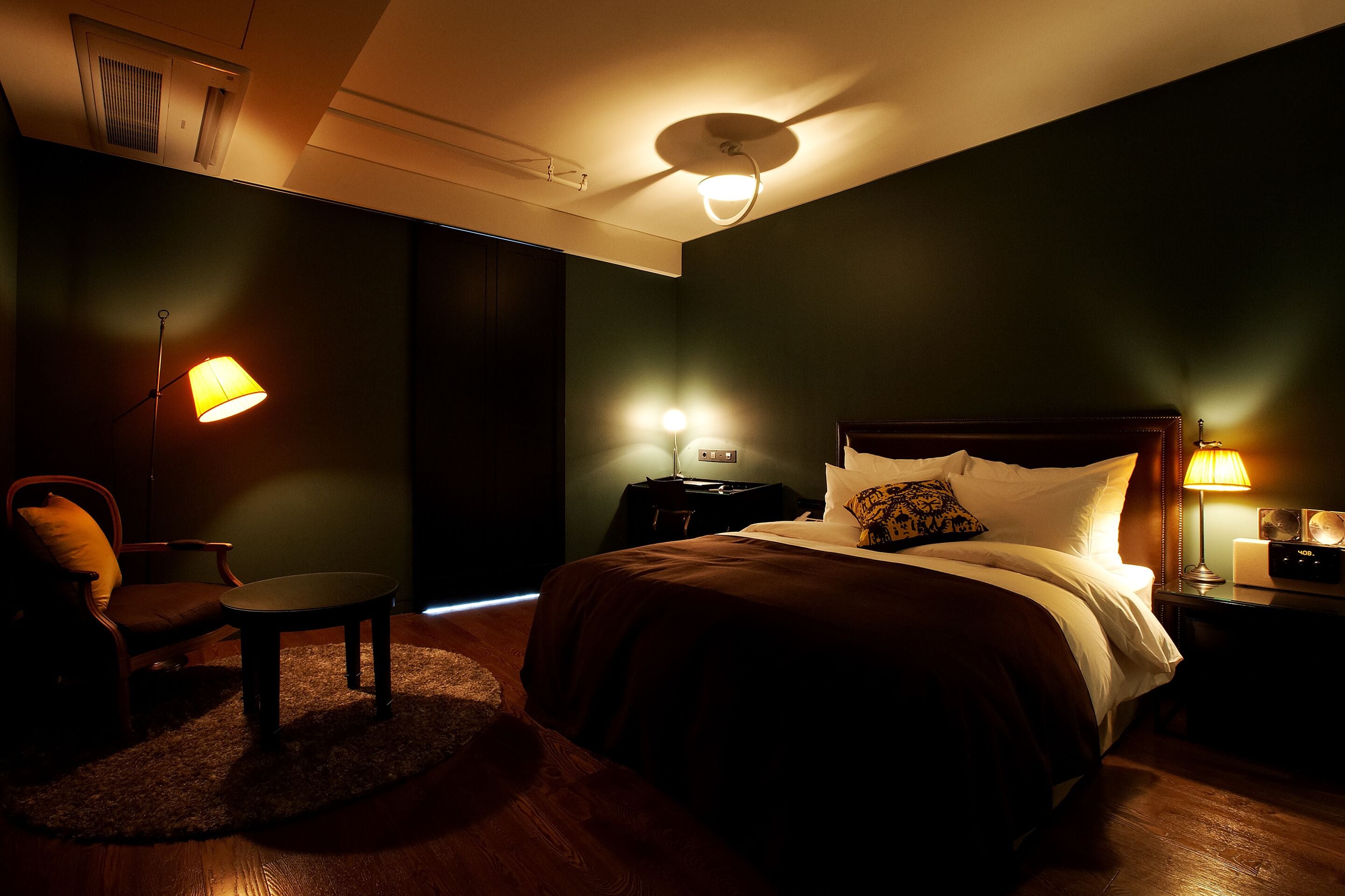 Un dormitorio tenuemente iluminado con una cama, una silla, una mesita y una mesita de noche con una lámpara.