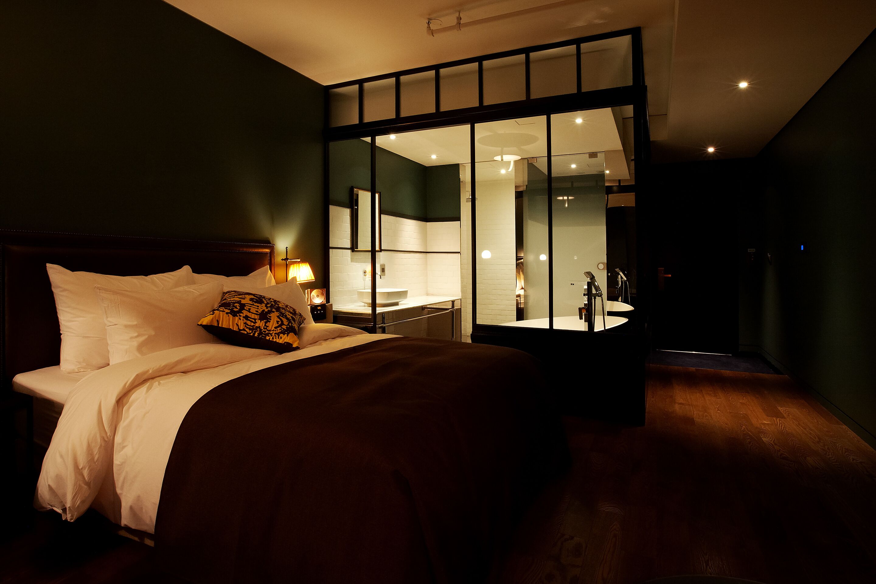 A modern hotel room with a large bed, a bedside table with a lamp, a glass partition wall, and a bathroom visible through the partition.
