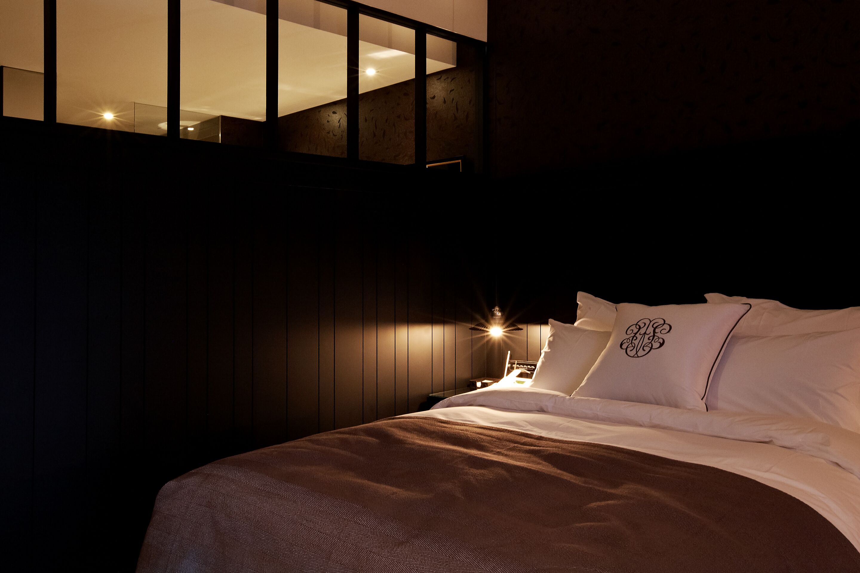 A dimly lit bedroom with a bed, a nightstand, and a window with a view of a building.