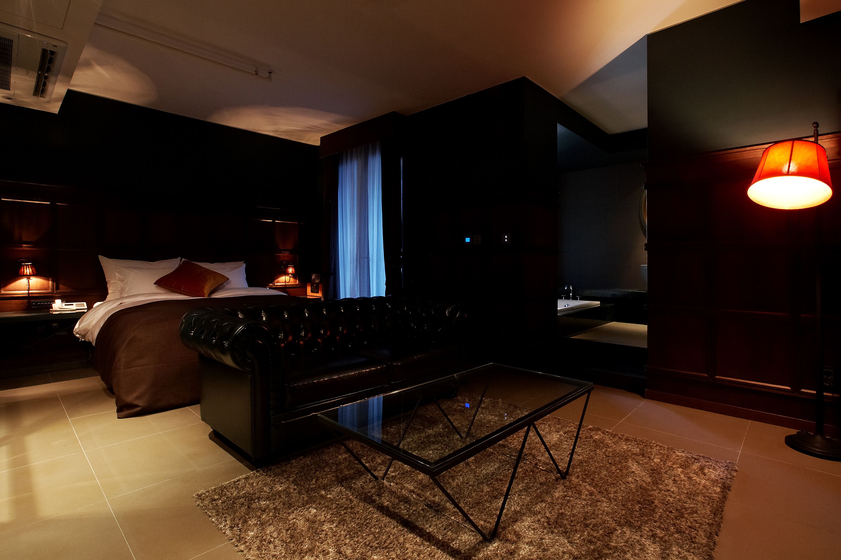 A modern hotel room with a bed, a sofa, a glass coffee table, and a large mirror.