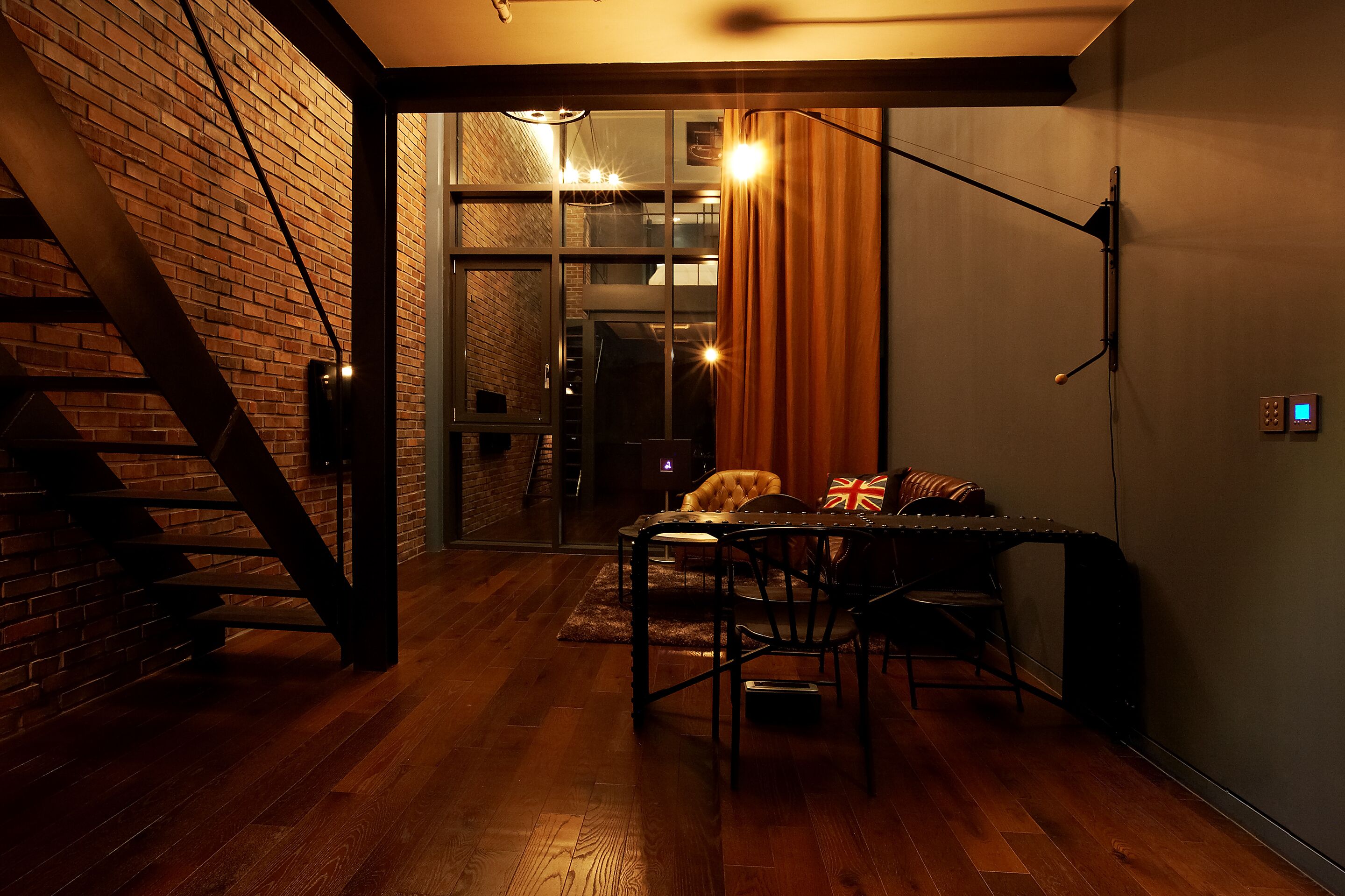 Une pièce chaleureuse avec un mur en briques, un plancher en bois et une table entourée de chaises. Un escalier se trouve à gauche et une fenêtre à droite.