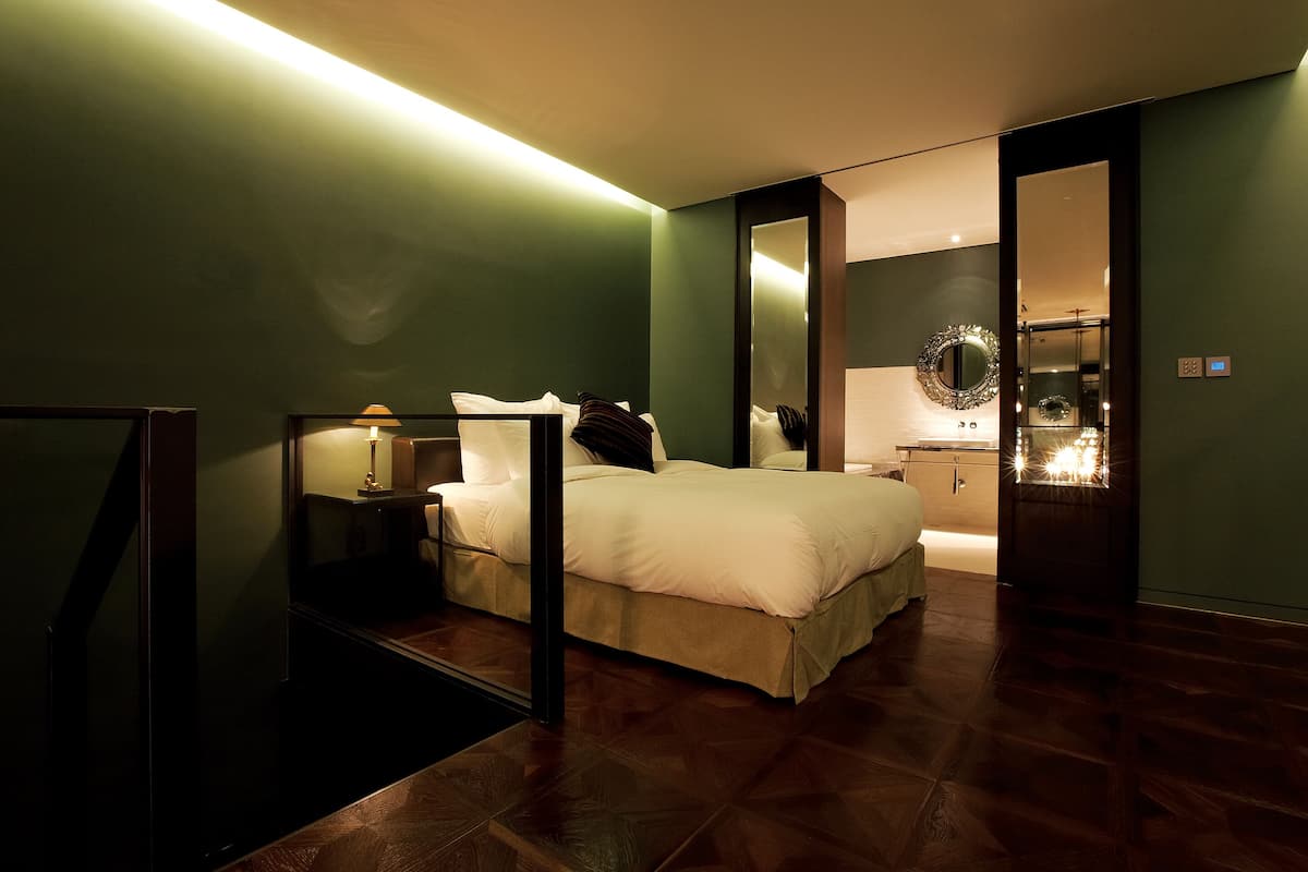 Una habitación de hotel moderna con una cama grande, una mesita de noche con lámpara, un espejo y vistas al baño.