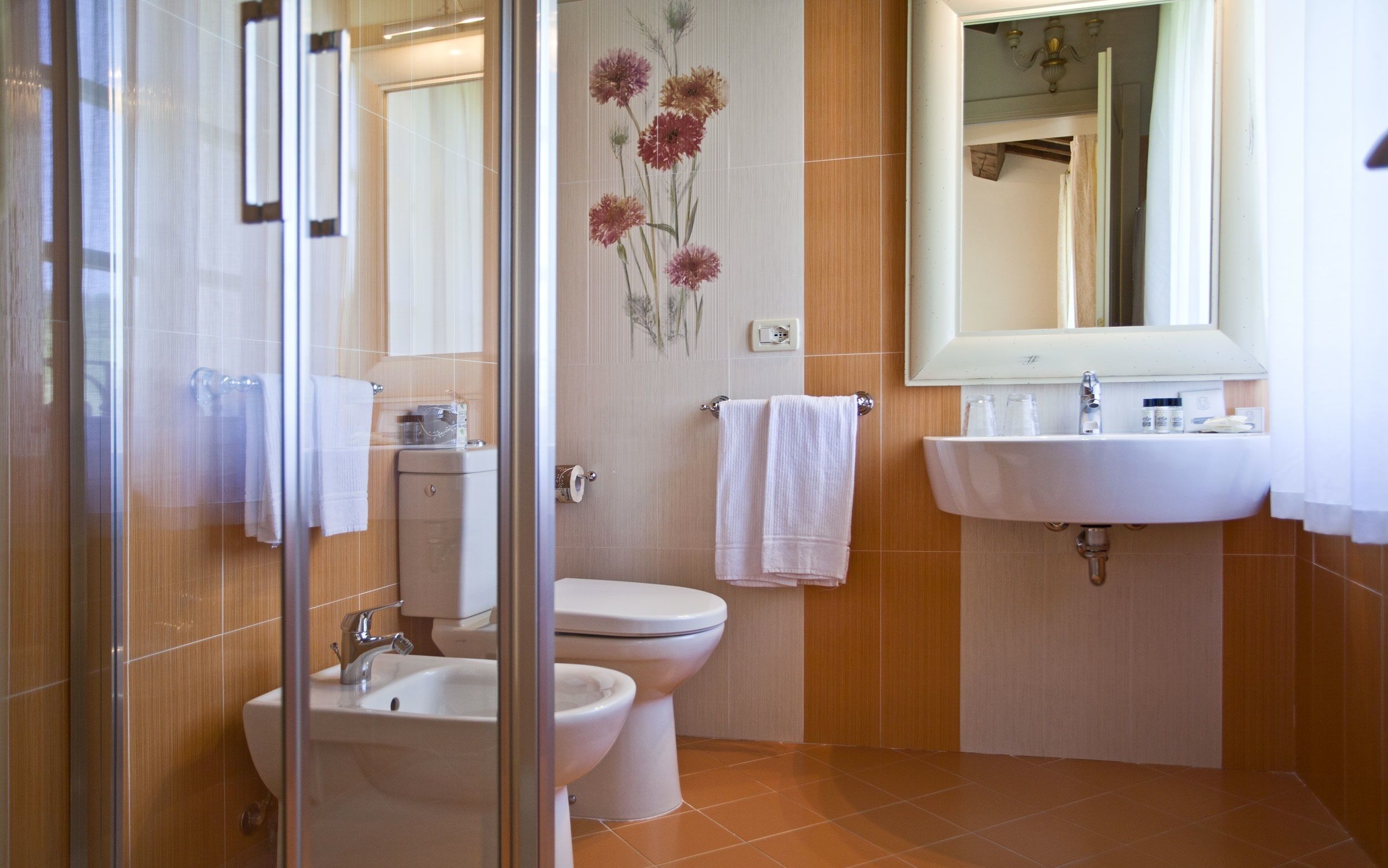 Un baño con inodoro, bidé y lavamanos. Hay un espejo sobre el lavamanos y un perchero con toallas.