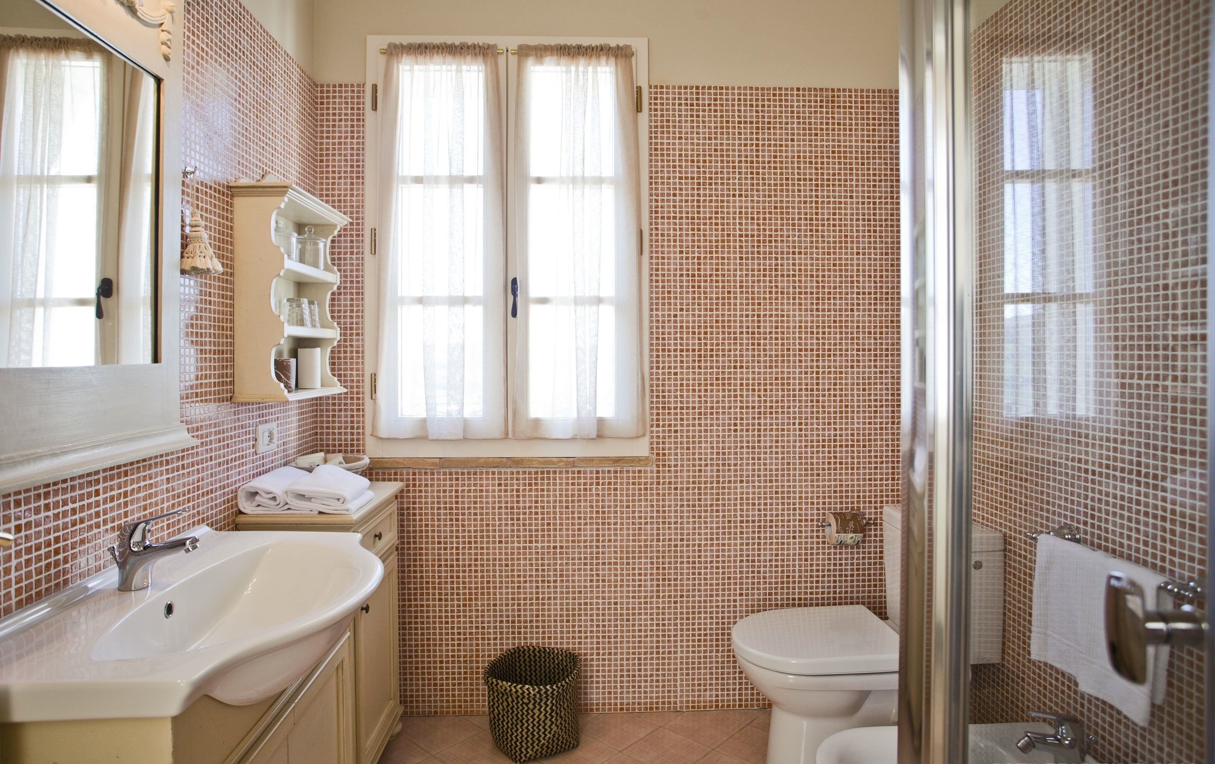 A bathroom with a sink, toilet, and shower. There is a window with curtains, a mirror, and a towel rack.