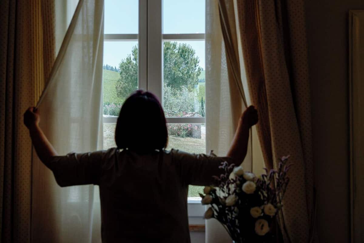 Une personne se tient près d’une fenêtre, avec des rideaux, un vase rempli de fleurs et une chaise.