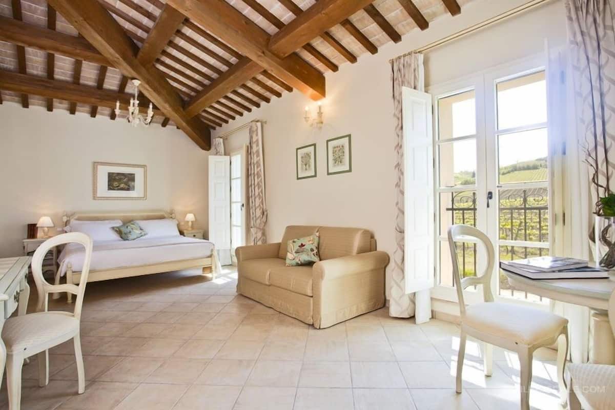 Une chambre à coucher avec un lit, un canapé, un bureau et des chaises. Il y a des fenêtres avec des rideaux et une vue sur l’extérieur.