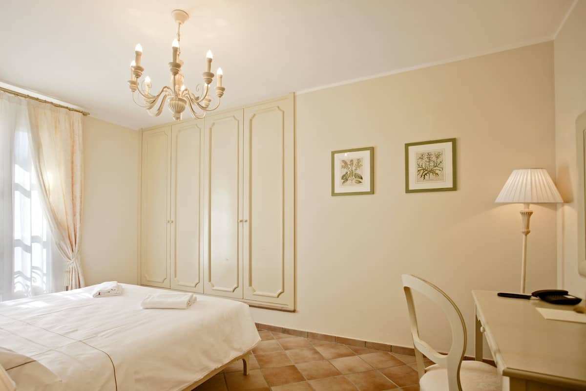 Une chambre d’hôtel comprenant un lit, un bureau, une chaise, une lampe et deux reproductions botaniques encadrées accrochées au mur.
