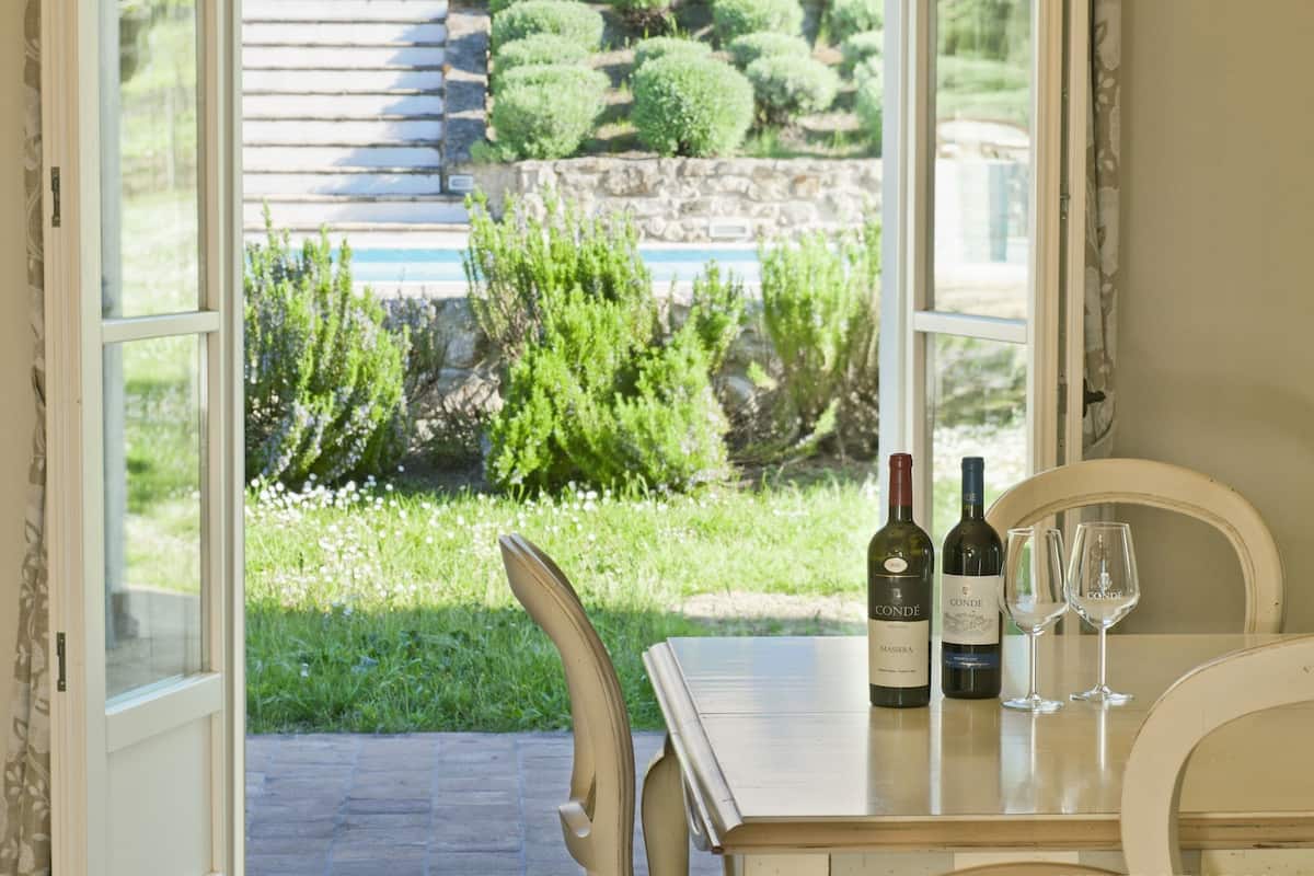 Une table avec deux bouteilles de vin et deux verres, placée près d’une fenêtre donnant sur un jardin avec un mur de pierre et de la verdure.