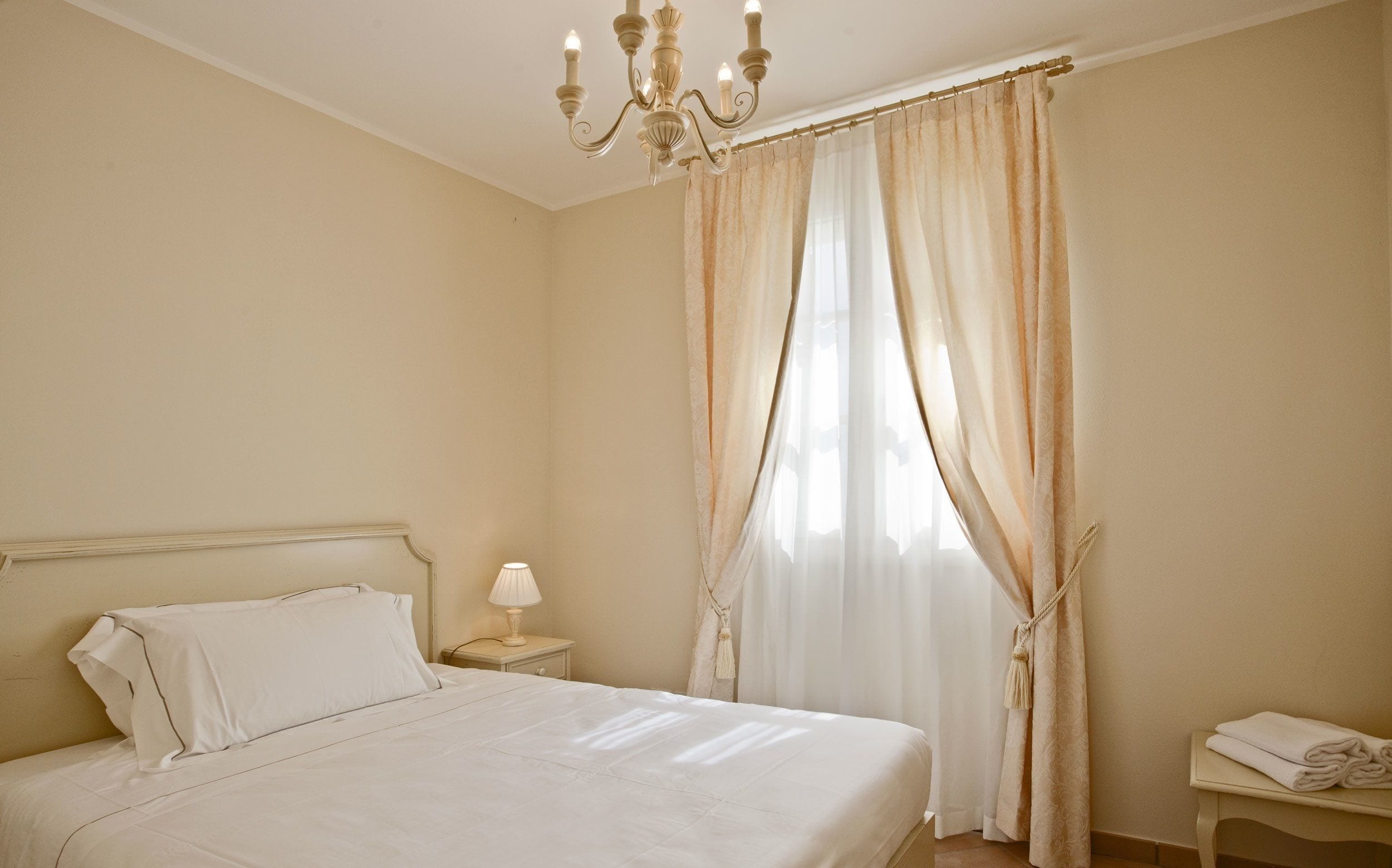 A neatly made bed with white linens, a bedside lamp, and a chandelier.
