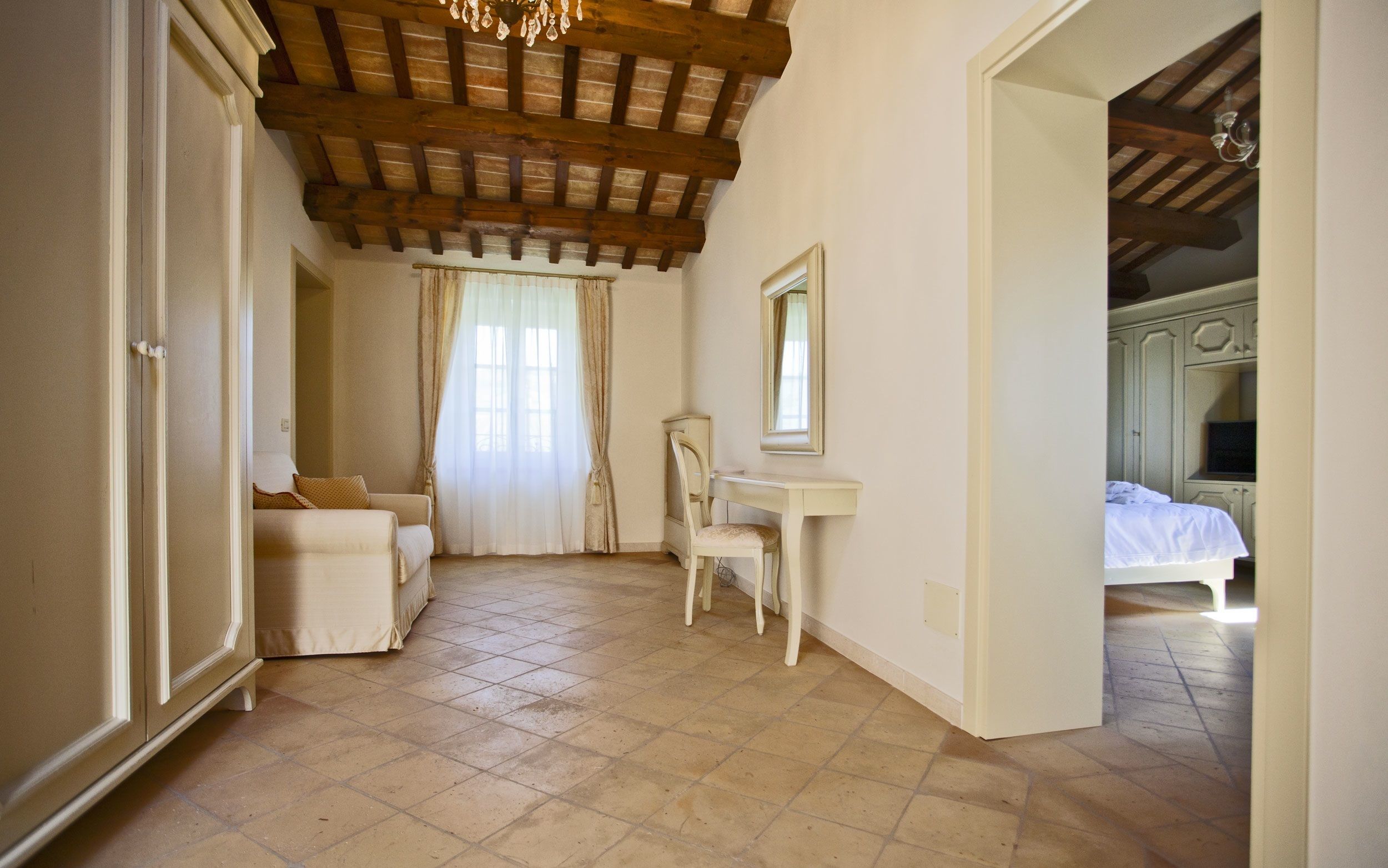 A bedroom with a bed, a desk, and a chair. There is a window with curtains and a chandelier.