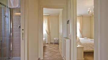 A hotel hallway leading to a bedroom with a bed, a nightstand, and a small table. There is a bathroom with a shower and a toilet visible through a glass door.