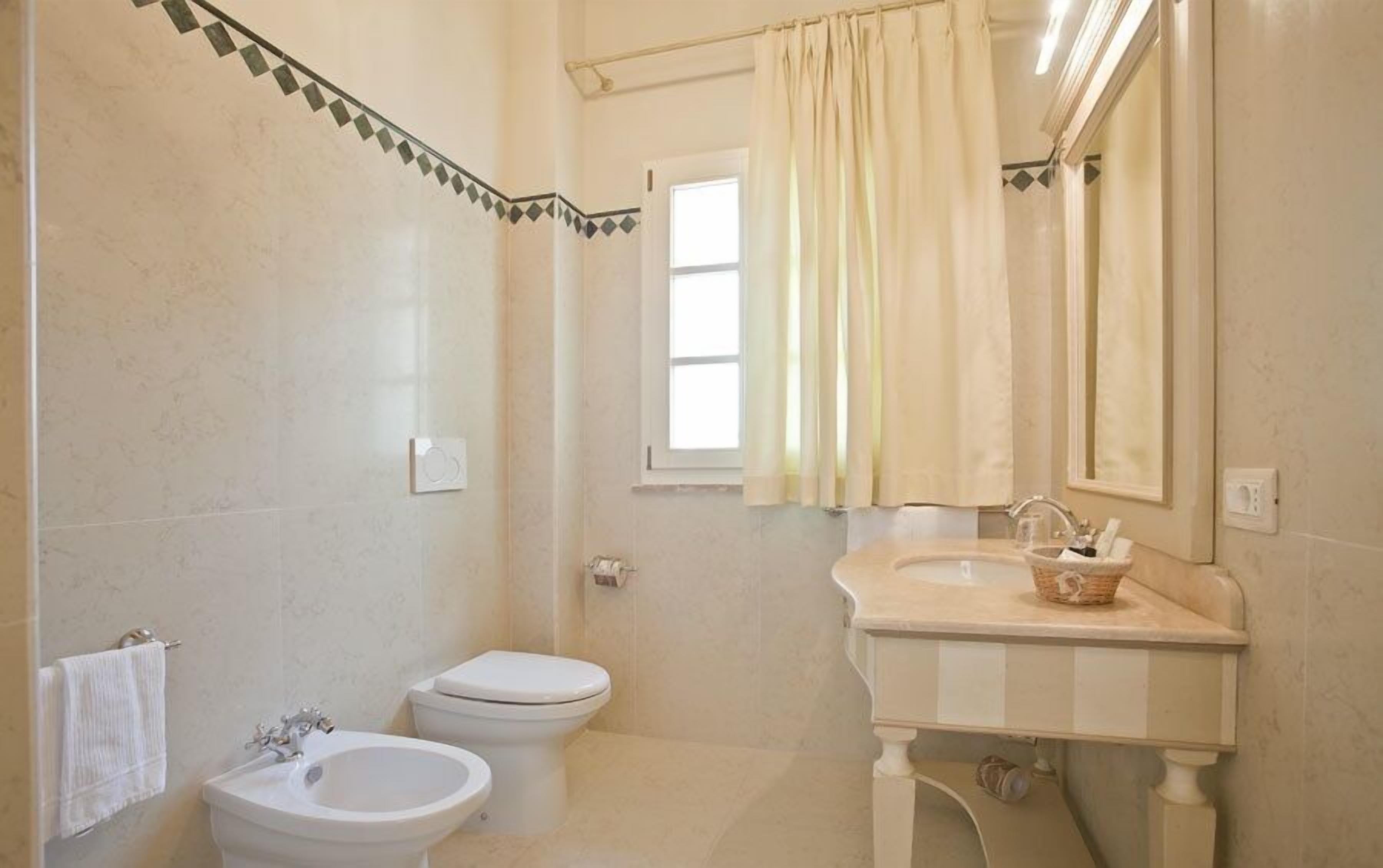 A bathroom with a toilet, bidet, sink, and a window with curtains.