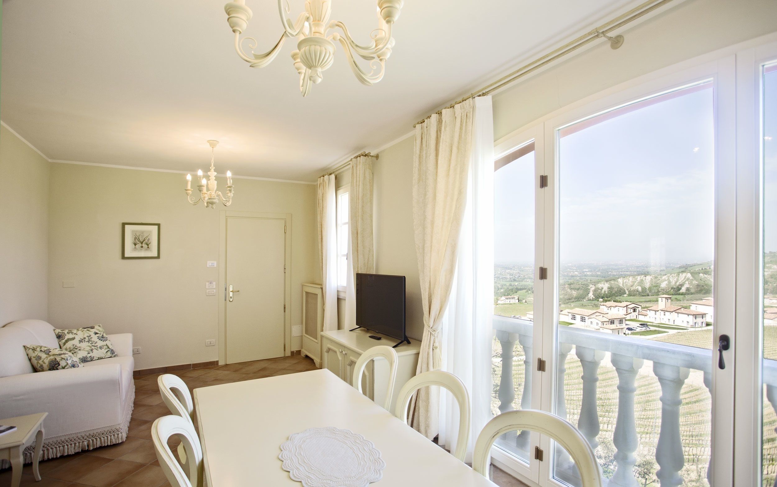 A room with a dining table, chairs, a television on a stand, a chandelier, a door, and a large window with a view of buildings and hills.