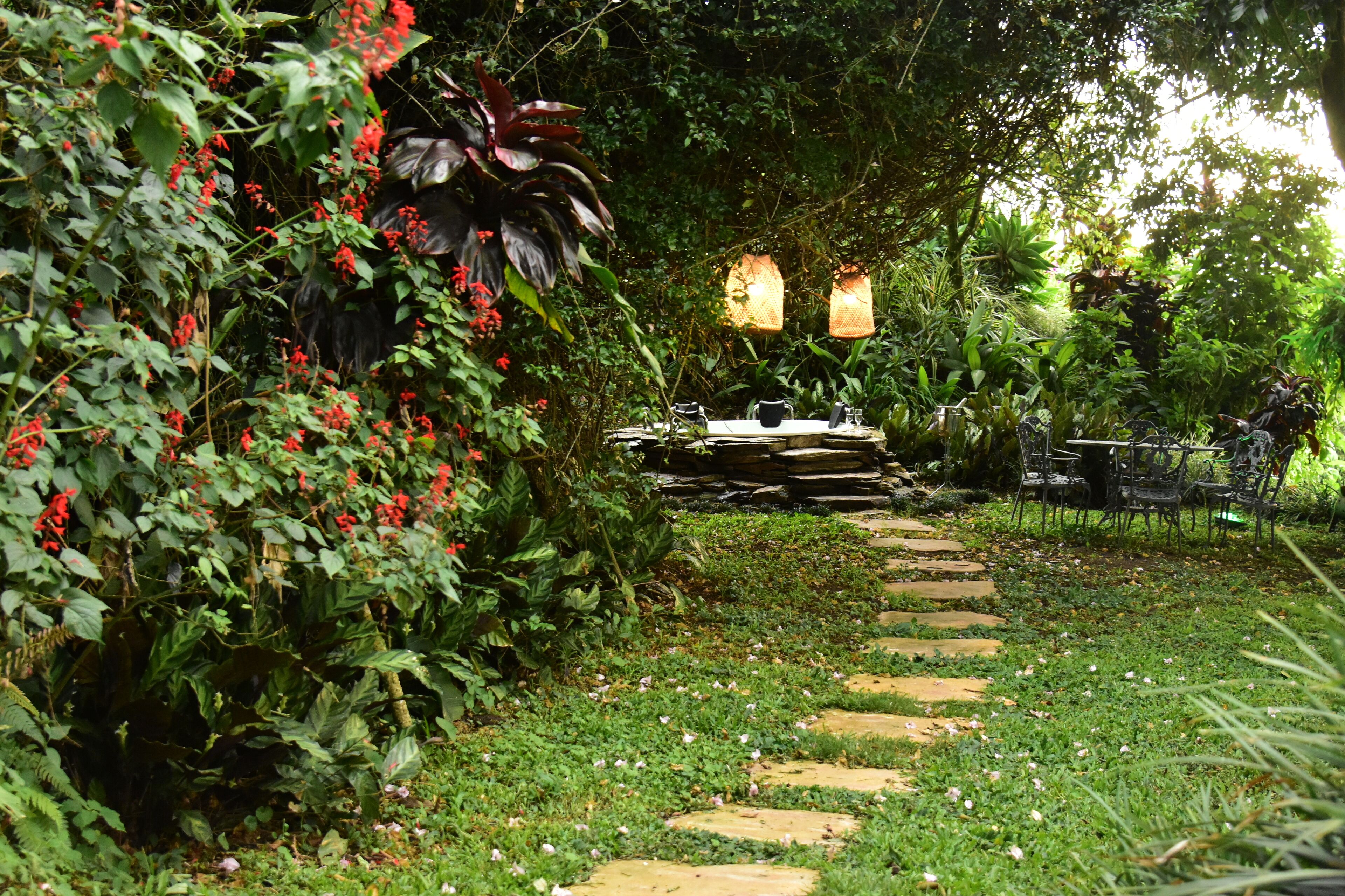 A garden pathway leading to a dining area with stone steps, surrounded by lush greenery and red flowers.