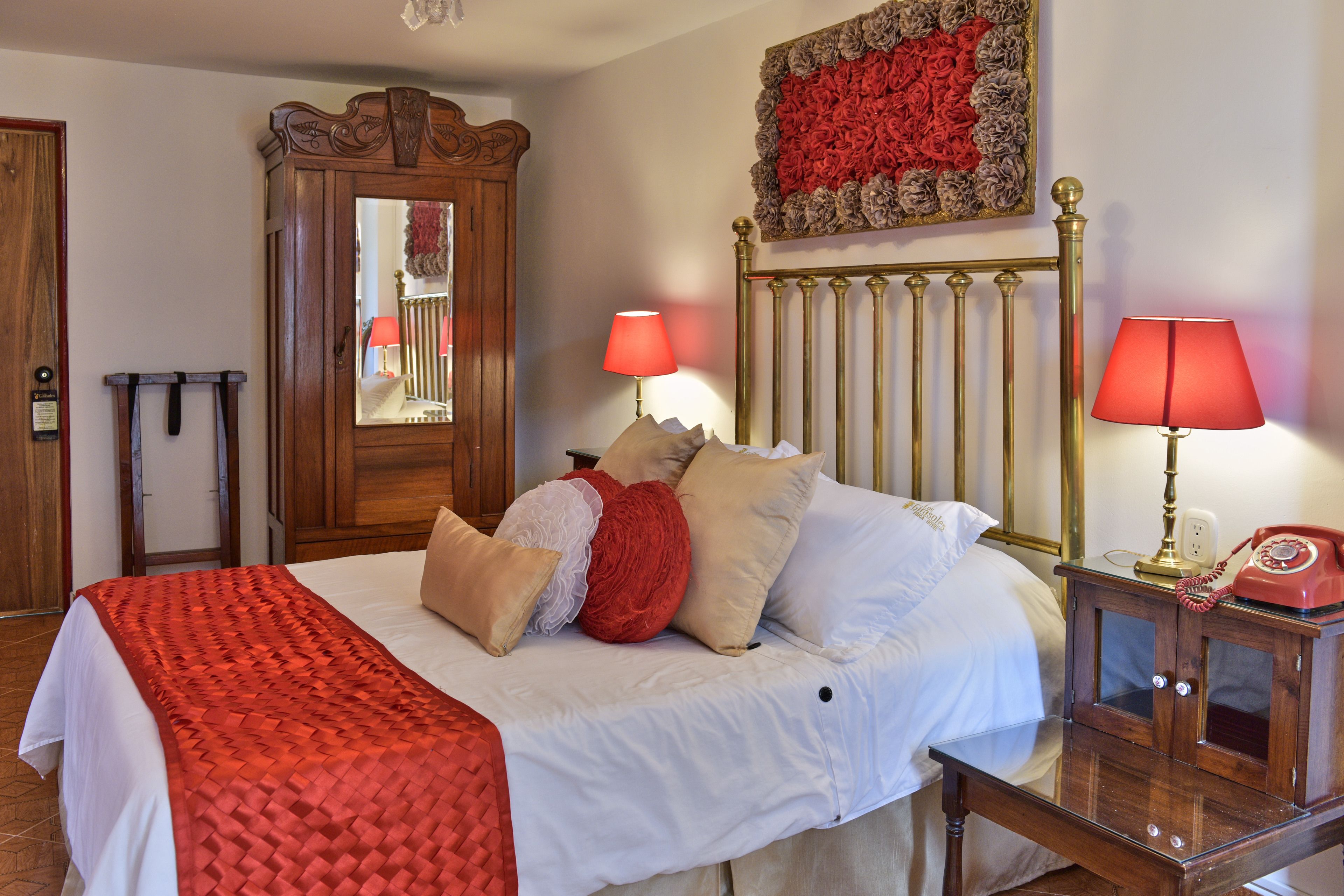 Un lit bien fait, recouvert d’une couverture rouge, avec deux lampes de chevet et un téléphone posé sur une table de nuit en bois.