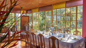 A dining area with a table set for a meal, surrounded by wooden chairs, large windows, and tropical plants. - Finca Hotel Los girasoles (Montenegro)