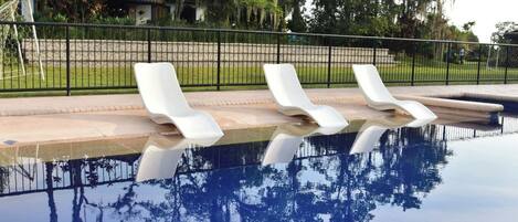 A swimming pool with three white lounge chairs, a fence, and a building with a tiled roof in the background.