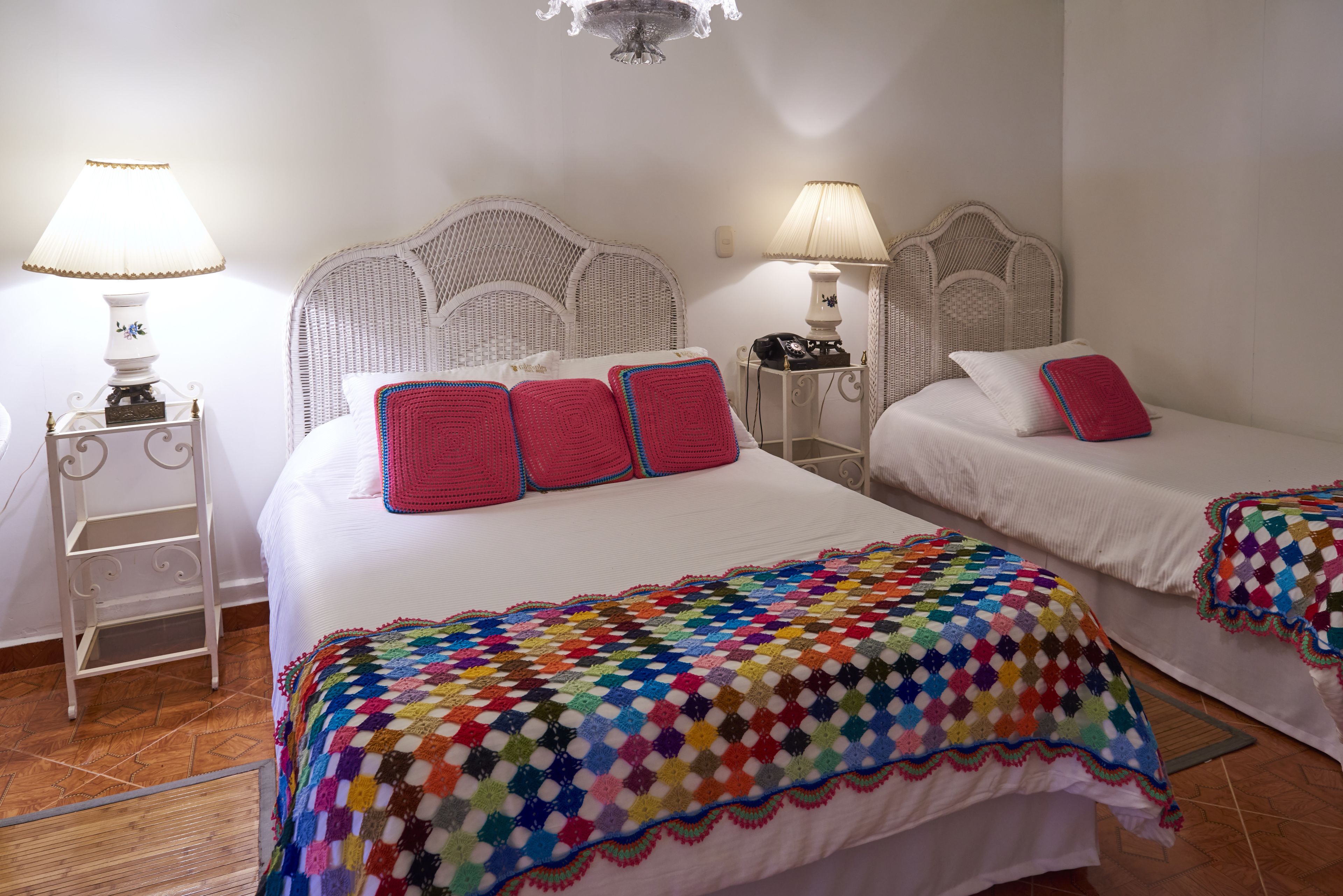 A hotel room with two beds, each with a white headboard, a colorful crocheted blanket, and a lamp on a bedside table.