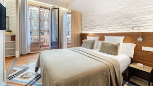 A modern hotel room with a large bed, a wooden headboard, a small bedside table, and a view of the city through a sliding glass door. - Fuster Apartments By Aspasios  (Barcelona)