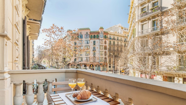 A balcony with a table set for breakfast, including croissants and orange juice, overlooking a city street with historic buildings. - Fuster Apartments By Aspasios  (Barcelona)